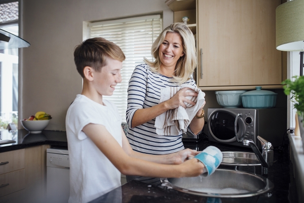 Mother and son doing the dishes together. They are talking and laughing as the boy washes and the mother dries dishes.