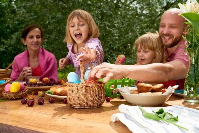 A mom and dad with two young daughters sitting down to an Easter meal outside. One of the girls is excitedly reaching to take the most beautiful Easter egg out of a basket on the table.