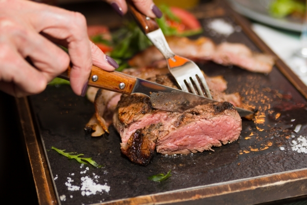 Using a fork and sharp knife, someone cuts into a juicy, medium-well steak on a cutting board.