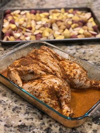 A seasoned roasting chicken with a pan of roasting potatoes in the background