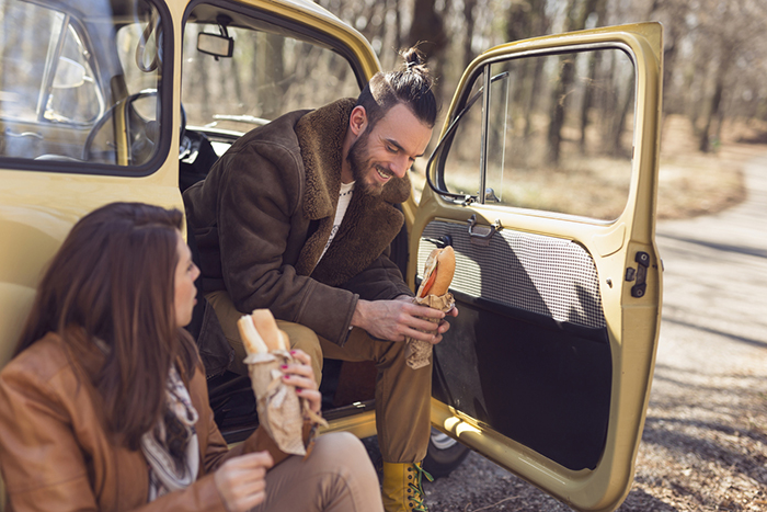 A man and woman take a lunch break with sandwiches outside their car on the side of the road